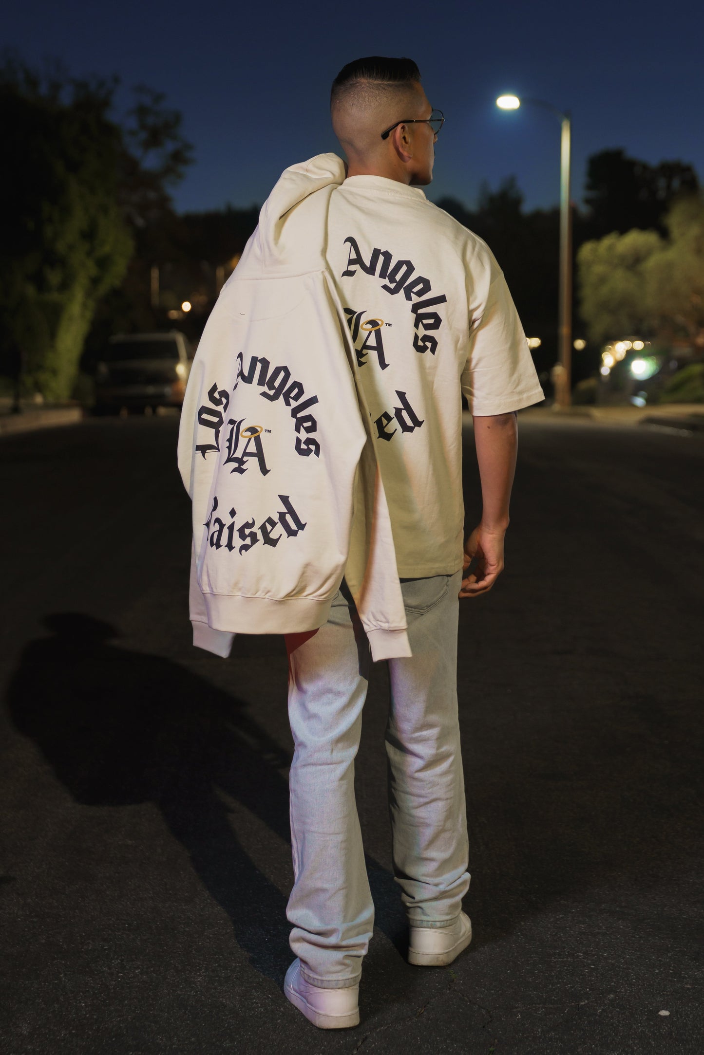 Person wearing a beige 'Los Angeles Raised' Shirt holding a beige Los Angeles Raised Sweater on a dark street.