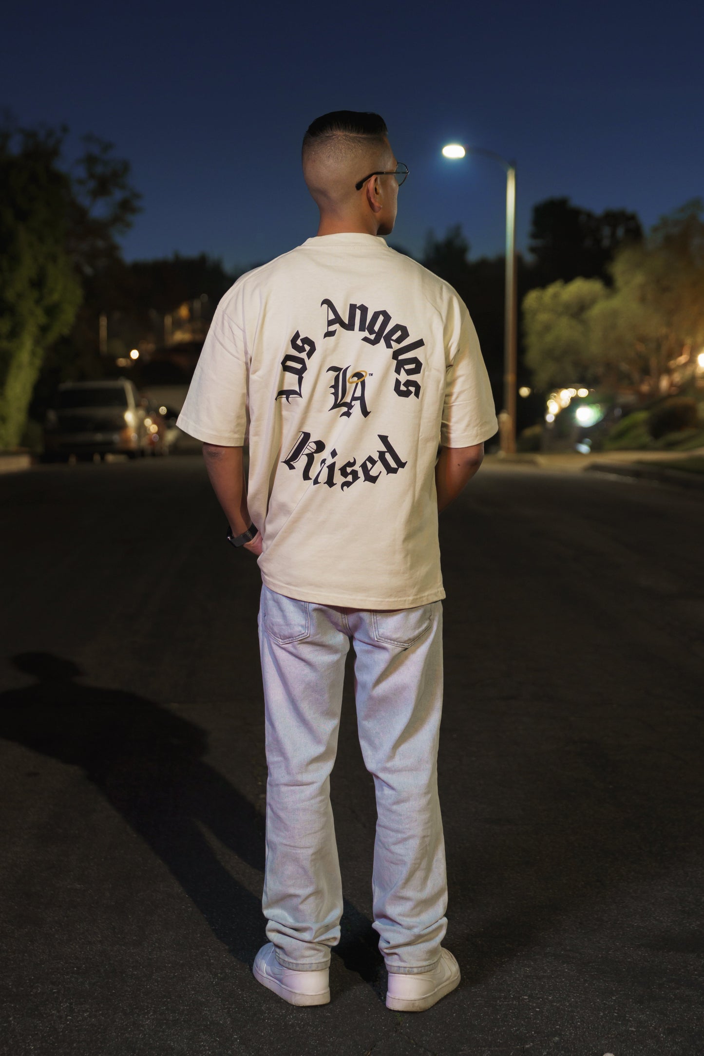 Person wearing a beige 'Los Angeles Raised' t-shirt standing on a street at night.